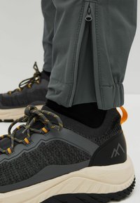 Gros plan sur une basket noire et grise avec des lacets soulignés de jaune, portée avec un pantalon gris doté d'une fermeture éclair à la cheville et d'un ourlet élastique.