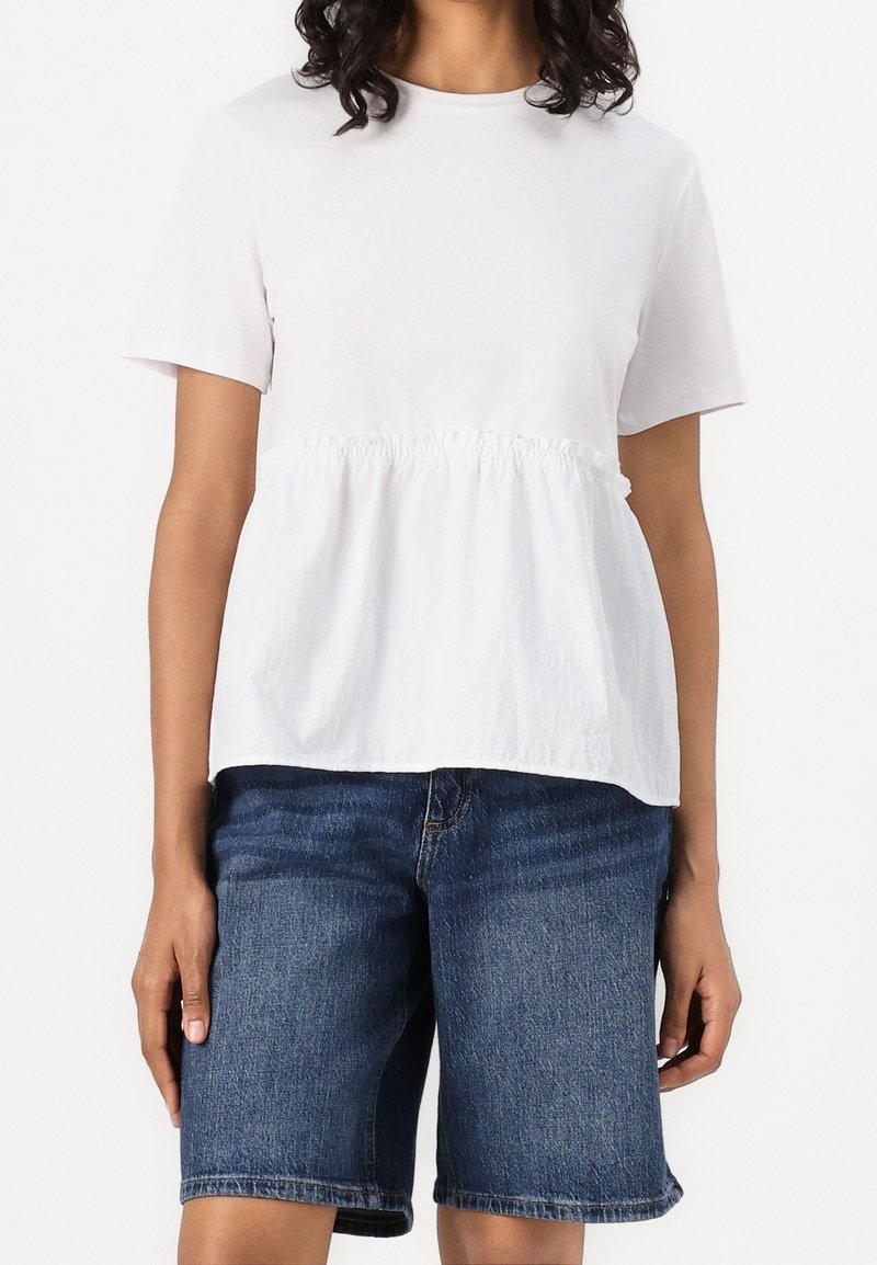 Personne portant un haut blanc à manches courtes avec un détail froncé à la taille et un short en jean bleu longueur genou, debout sur un fond blanc.