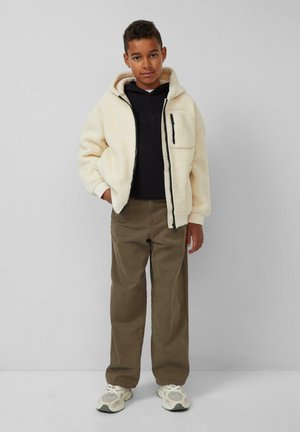 Jongen in een beige fleecejack over een zwarte hoodie, met bruine broek en witte sneakers, staand tegen een effen lichtgrijze muur.