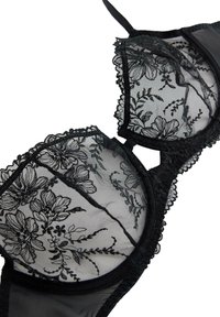 Reggiseno in pizzo nero con ricami floreali, coppe trasparenti e bordi smerlati. Il design include spalline regolabili e un sottostrato in raso liscio.