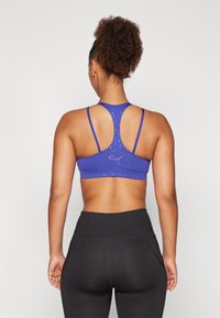 Paarse sportbh met racerback ontwerp, voorzien van een gespikkeld patroon en een klein logo op de achterkant, gecombineerd met hoge zwarte leggings.