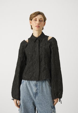 Black blouse with textured floral design, open shoulder cutouts, collared neckline, and long sleeves. Worn with distressed denim shorts.