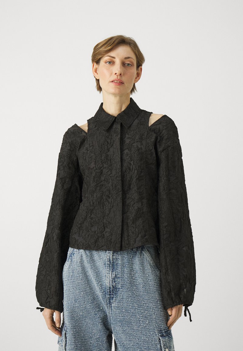 Black blouse with textured floral design, open shoulder cutouts, collared neckline, and long sleeves. Worn with distressed denim shorts.