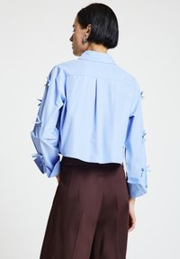 Donna che indossa una blusa corta azzurro chiaro con dettagli a fiocco sulle maniche e pantaloni scuri a vita alta, mostrata di spalle su uno sfondo bianco.