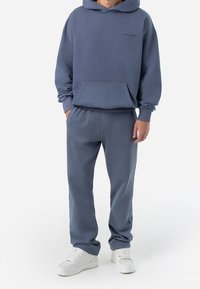 Sudadera azul con bolsillo delantero y capucha con cordón, combinada con pantalones deportivos azules a juego. Zapatillas blancas completan el conjunto.