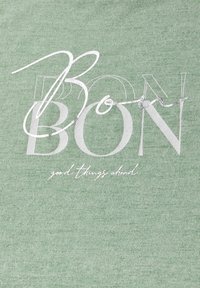 Světle zelená tkanina se vzorem obsahujícím slovo "BON" v bílé barvě s překrytem v písmu, a frázi "dobré věci před námi" v jemném písmu níže.
