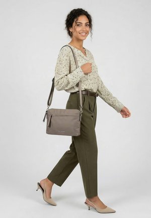 Frau läuft, trägt ein gemustertes langärmeliges Oberteil, olivgrüne Hose, beige Absätze und eine graue Umhängetasche auf der Schulter.