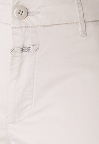 Gros plan sur la ceinture beige clair d’un pantalon avec un bouton, une passant de ceinture, une poche avant et une petite étiquette en tissu cousue.
