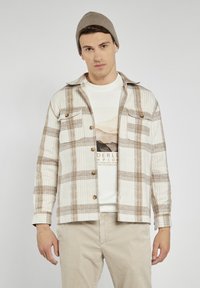 Koszula flanelowa w kolorze kremowym z brązowym wzorem w kratkę, z dwiema kieszeniami na piersi i brązowymi guzikami. Nosi się ją na jasnym t-shircie z grafiką.