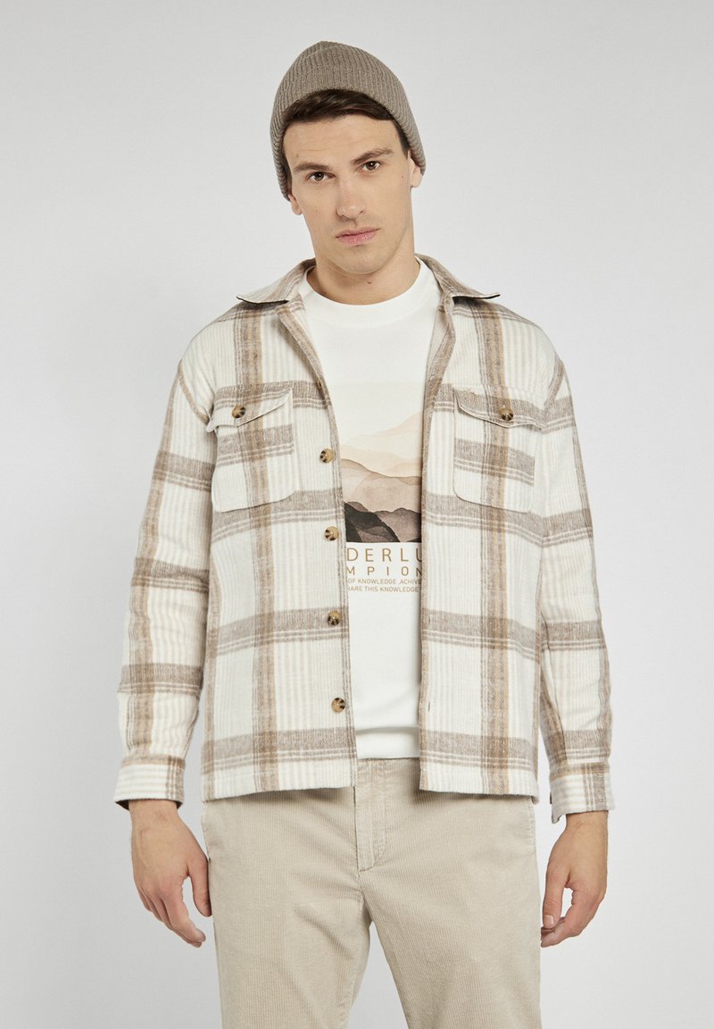 Koszula flanelowa w kolorze kremowym z brązowym wzorem w kratkę, z dwiema kieszeniami na piersi i brązowymi guzikami. Nosi się ją na jasnym t-shircie z grafiką.