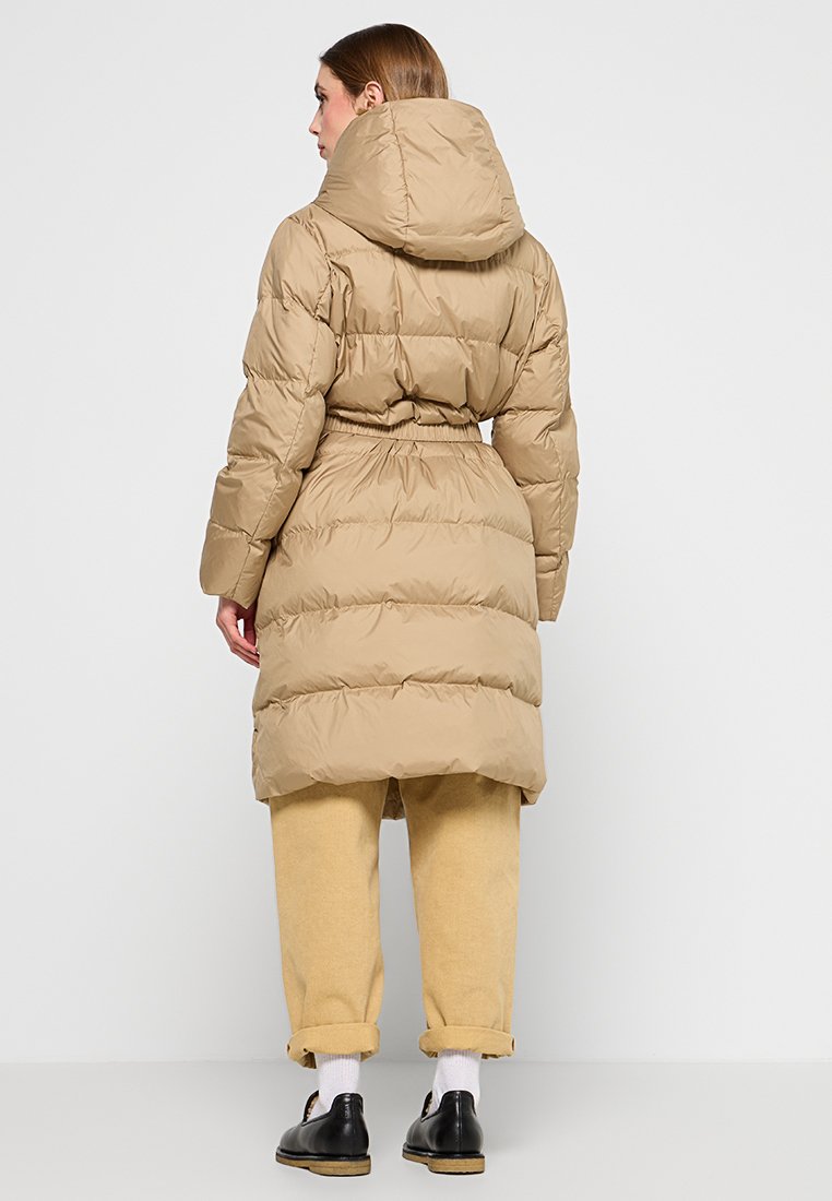 Cappotto puffo beige con cappuccio, caratterizzato da una vita sfiancata, texture trapuntata e orlo a lunghezza ginocchio. Indossato sopra pantaloni e scarpe di colore chiaro.