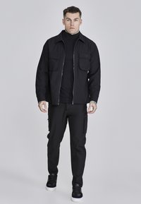 Chaqueta negra con cremallera y dos bolsillos en el pecho, superpuesta a una parte superior negra de cuello alto, combinada con pantalones cargo negros y zapatillas negras.