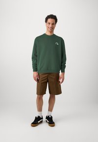 Sweatshirt en coton vert avec des poignets et un col côtelés, associé à un short marron et des baskets noires avec des accents blancs et des semelles en caoutchouc.