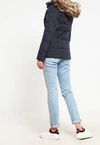 Marineblå dunjakke med pelsforet hætte, med quiltet design. Båret med lyseblå jeans og hvide sneakers.