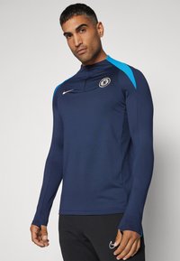Maglietta sportiva a maniche lunghe di colore blu navy con mezzo zip al collo, dettagli blu sulle spalle e un logo circolare sul petto. Presenta una vestibilità slim.