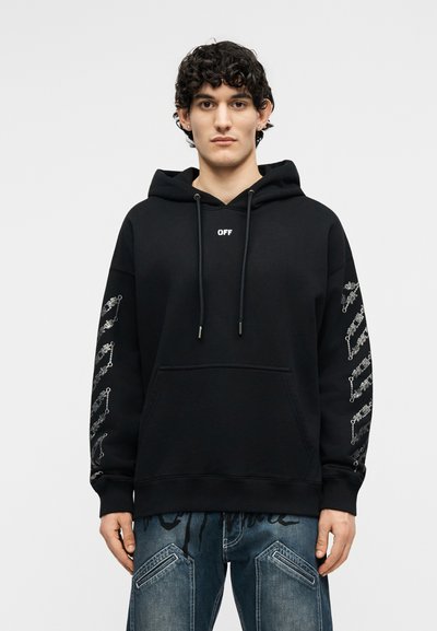 Jauns vīrietis ar cirtainiem matiem, valkājot melnu kapuci ar uzrakstu "OFF" priekšpusē un baltām ķēžu rakstu dizainiem uz piedurknēm, kopā ar zilām džinsu biksēm.