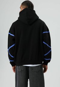 Zwarte hoodie met blauwe gebogen accenten op de mouwen, gemaakt van zacht materiaal. Voorzien van een grote capuchon en verlaagde schouders voor een ontspannen pasvorm.