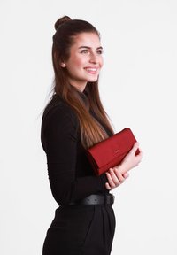 Red fabric wallet with a smooth texture, featuring a folded design and an embossed logo, held by a person in a black top and pants.