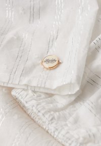 Hvid tekstureret stof med sølv lodrette striber og et guldindrammet rundt mærke, der er indgraveret med "Superdry" i kursiv.