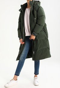 Długa zielona pikowana kurtka z kapturem, zamkiem błyskawicznym i zatrzaskami, bocznymi kieszeniami oraz ściągaczami na rękawach, noszona na jasnoróżowej bluzce i niebieskich dżinsach.