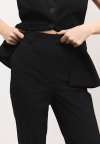 Pantaloni di un completo nero su misura con un design elegante, parte anteriore piatta e tasche laterali. Il tessuto appare liscio con una vestibilità strutturata.