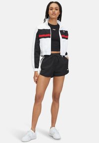 Geknipte witte jas met zwart en rode strepen over een zwarte crop top, gecombineerd met zwarte shorts en witte sneakers.