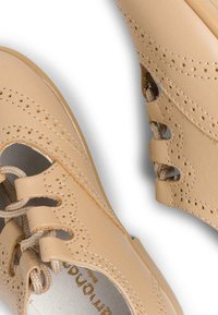 Beige kožené boty s kulatou špičkou, zdobené perforovanými detaily, šněrováním a vystřihovanými akcenty po stranách. Hladký povrch a minimalistický design.