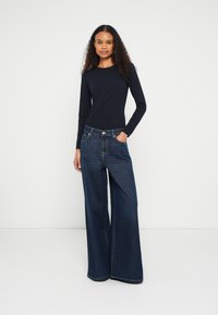 Mulher de cabelo encaracolado a usar uma camisola de manga comprida preta e jeans azul-escuro de pernas largas, em pé contra um fundo branco liso.