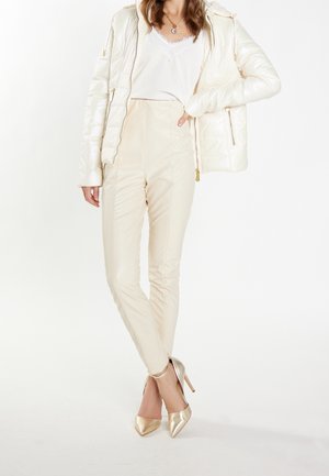 Mujer con chaqueta acolchada color crema sobre una blusa blanca con encaje, pantalones beige de cintura alta y tacones dorados puntiagudos, de pie con las piernas cruzadas.