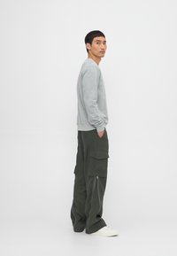Gray sweatshirt with long sleeves, paired with dark green cargo pants featuring pockets and zippers. Model stands side view against a white background.