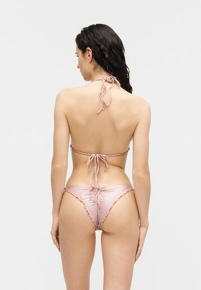 Hosszú, sötét, nedves hajjal rendelkező nő világos rózsaszín, redős bikiniben, háttal áll a fényképezőgép felé egy egyszínű világos háttér előtt.