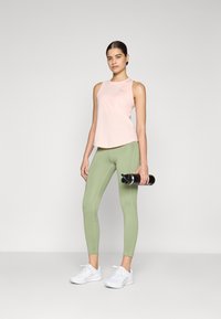 Šviesiai rožinė be rankovių palaidinė, žaliasias leggingai ir baltos sportinės bateliai. Modelis laiko juodą vandens butelį, fone - neutrali aplinka.
