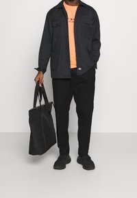 Camisa negra de botones sobre una camiseta gráfica naranja, combinada con pantalones negros y un bolso acolchado negro. Zapatillas negras completan el look.