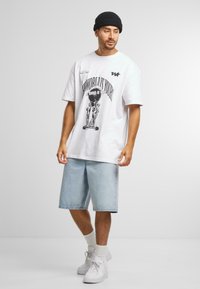 Hvitt t-skjorte med svart grafisk trykk som viser en figur, kombinert med lyseblå denimshorts. Brukt med hvite sneakers og en svart beanie.