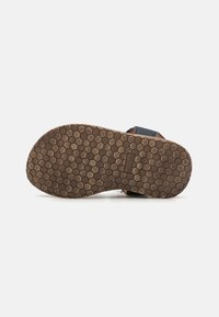 Brun gummiyttersåle med sekskantet mønster; designet for grep. Har blå stropper og lærdetaljer. Flat, åpen stil for pusteevne.