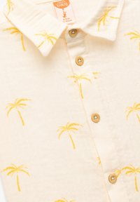 Crèmekleurig babyshirt met gele palmbomenpatronen, een kraag en vier bruine knopen aan de voorkant.