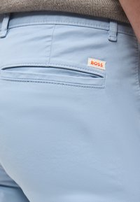 Πίσω τσέπη σε ανοιχτό μπλε παντελόνι με μικρή λευκή ετικέτα που γράφει "BOSS", φορεμένο με μπεζ πλεκτό πουλόβερ.