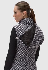 Pūkajamā veste ar melnbalto houndstooth rakstu, ar mākslīgās ādas oderi apņemtu kapuci un kniedei. Izgatavota no viegla auduma.