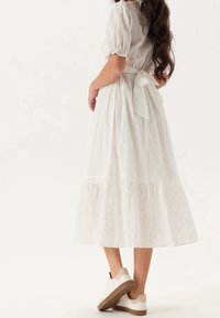 Biała, warstwowa sukienka midi z haftowanymi kwiatowymi wzorami, związaną w talii, z krótkimi bufkami, zestawiona z białymi sneakersami.