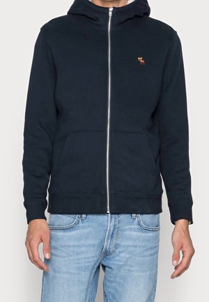 Mężczyzna w czarnej bluzie na zamek z małym pomarańczowym logo łosia na piersi i jasnoniebieskich dżinsach, stoi ze swobodnie opuszczonymi ramionami.