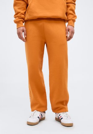 Homme portant un pantalon de survêtement orange et un sweat à capuche assorti, debout, montrant des baskets blanches avec des rayures rouges et bleues.