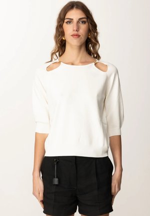 Maglione bianco a maniche corte con dettagli sulle spalle a taglio, rifiniture dorate e una trama a maglia morbida. Abbinato a pantaloni corti neri.