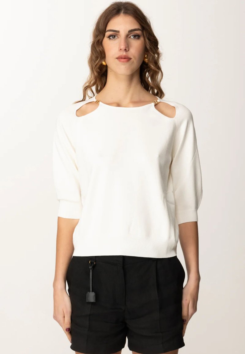 Maglione bianco a maniche corte con dettagli sulle spalle a taglio, rifiniture dorate e una trama a maglia morbida. Abbinato a pantaloni corti neri.