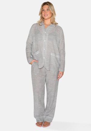 Jongevrouw staat blootsvoets, draagt een grijs-wit gestreept pyjamashirt met lange mouwen en bijpassende broek, glimlacht licht.