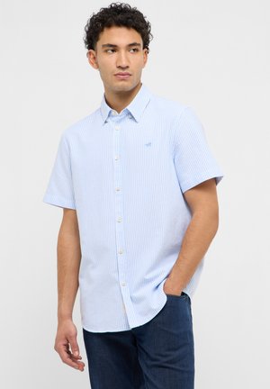 Mężczyzna w jasno-niebiesko-białej, paskowanej, krótkorękawowej koszuli z kołnierzykiem, ciemnoniebieskich dżinsach, z ręką w kieszeni, patrzący w lewo.