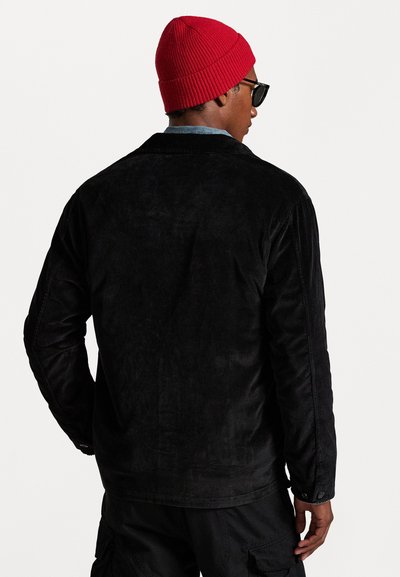 Black corduroy jacket with long sleeves, featuring a textured surface. Worn over a denim shirt, paired with a red knit beanie and sunglasses.