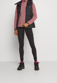 Doudoune sans manches noire avec capuche portée sur un haut à col montant rayé rouge et blanc, associée à des leggings noirs et des bottes noires avec des accents roses.