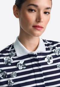 Polo de rayas navy y blancas con cuello blanco, adornado con múltiples apliques florales plateados texturizados y lentejuelas en el pecho.