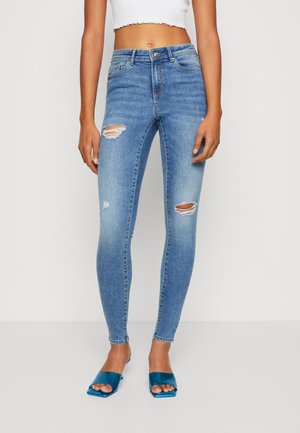 Femme portant un jean skinny déchiré bleu clair taille haute, un top court blanc et des sandales à talons ouvertes bleues, se tenant devant un fond uni.
