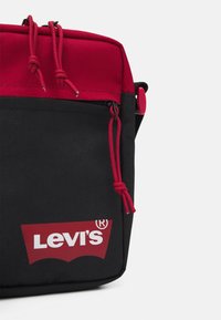 Borsa a tracolla nera in tessuto con parte superiore rossa, chiusura a cordoncino e logo rosso di Levi's ben visibile sul fronte. Presenta una tasca con zip.